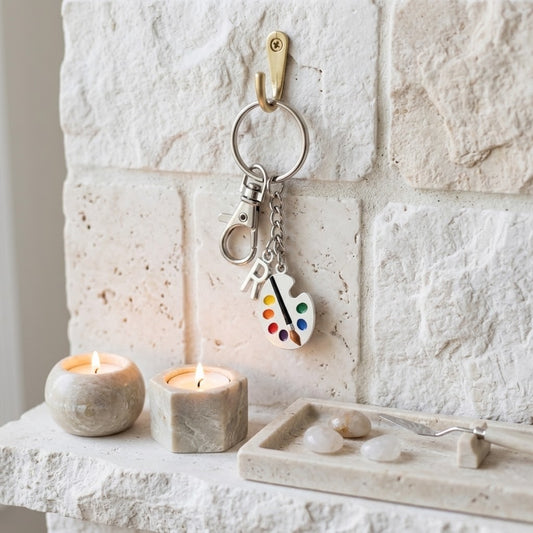 A silver keychain with a paint palette charm hangs from a brass wall hook on a white stone wall above a marble mantel with two lit candles and a crystal tray.