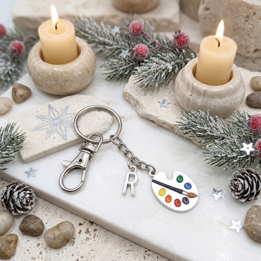 A silver keychain with a lobster clasp, a hanging letter 'R' charm, and an artist paint palette charm, styled on a white stone tray with candles, pine branches, and small stones.
