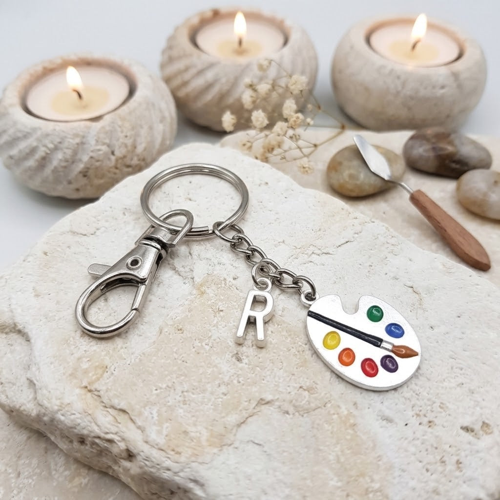 A silver metal keychain with a lobster clasp, a hanging letter 'R' charm, and a paint palette charm filled with colourful paint dots and a paintbrush, displayed on a textured stone with candles and small props in the background.