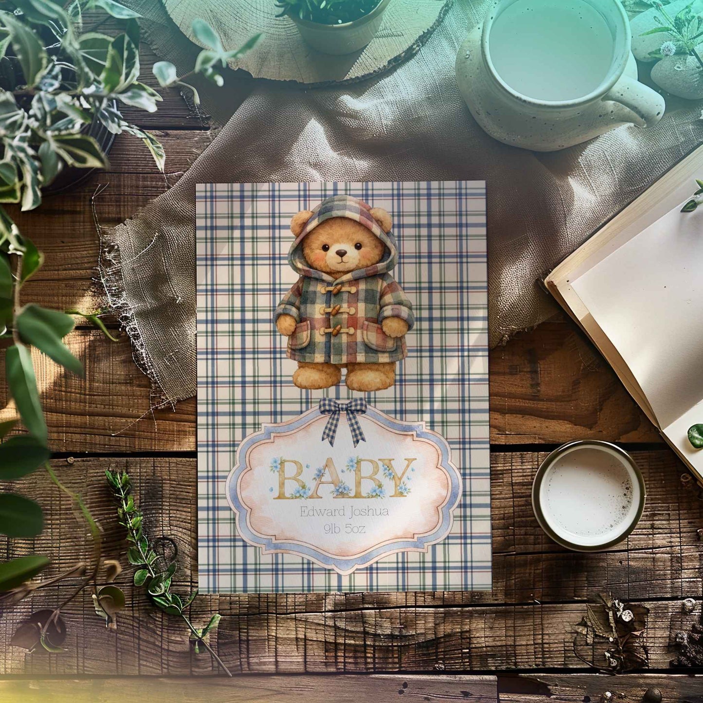 A baby shower card featuring a teddy bear in a plaid hooded jacket, with a decorative label reading 'BABY', styled on a rustic wooden table with plants, a mug, and an open book.