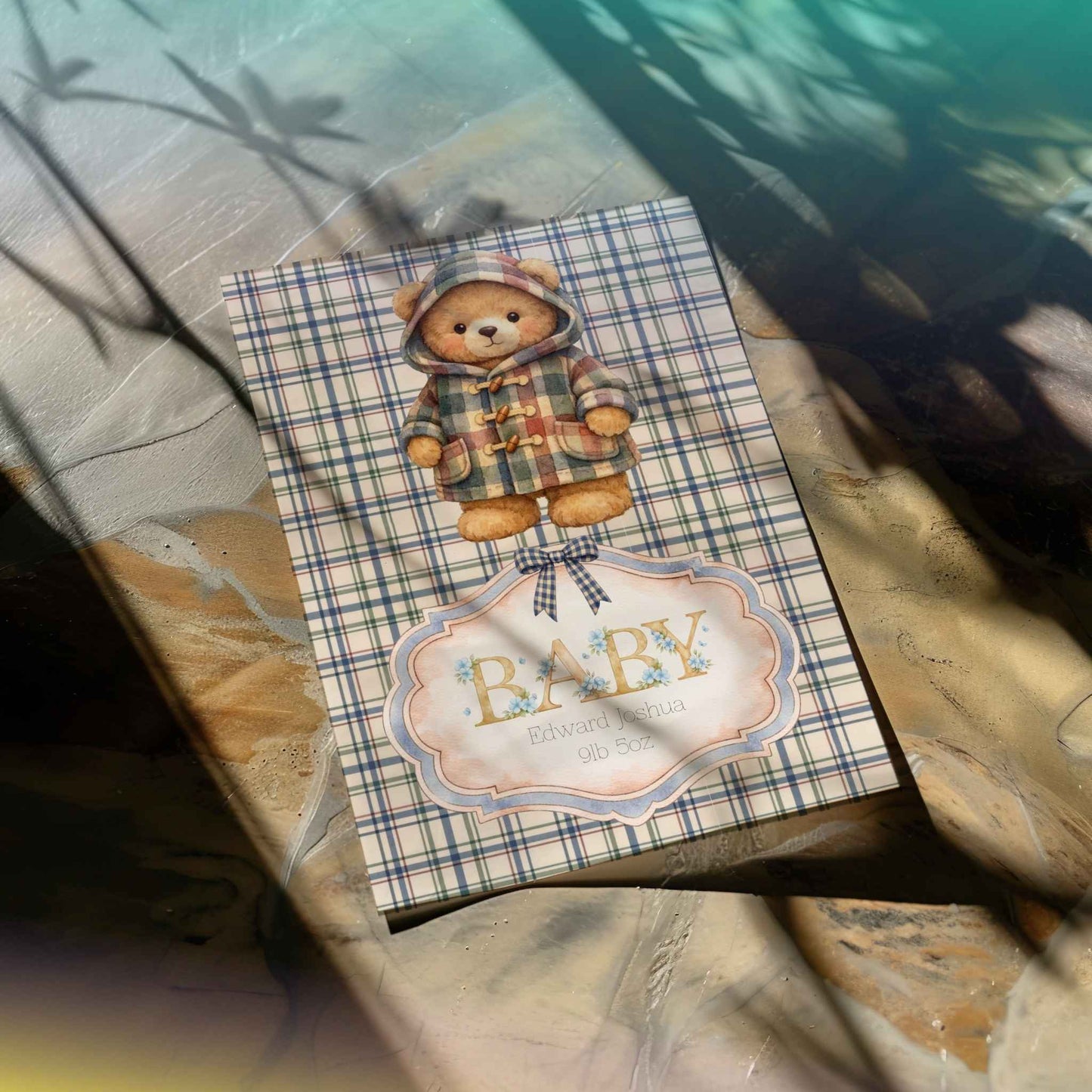 A baby shower card featuring a teddy bear in a plaid hooded jacket, with a decorative label reading 'BABY' and baby's name and birth weight, photographed on a stone surface with dappled sunlight and shadows.