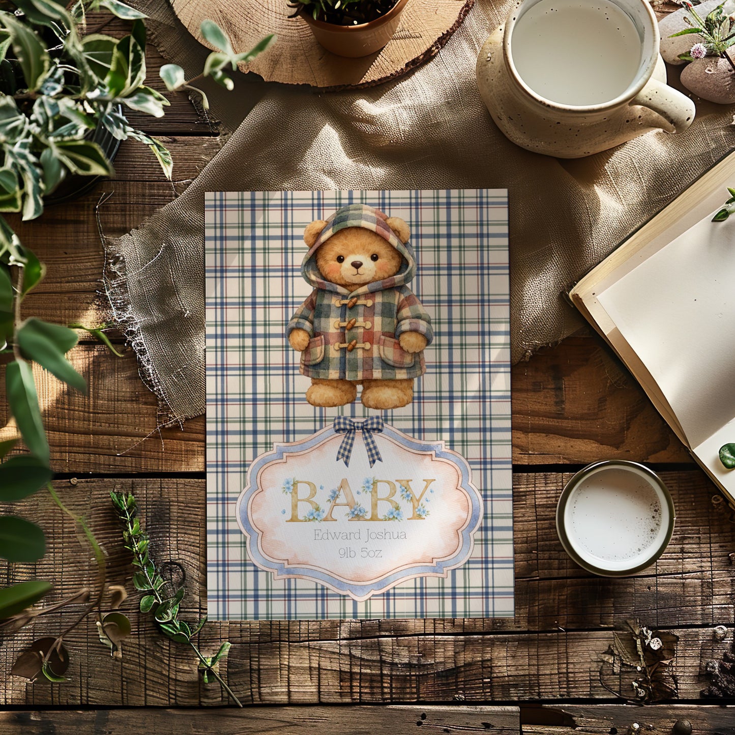 A baby shower card featuring a teddy bear in a plaid hooded jacket, styled on a rustic wooden table with a mug, book, and plants.