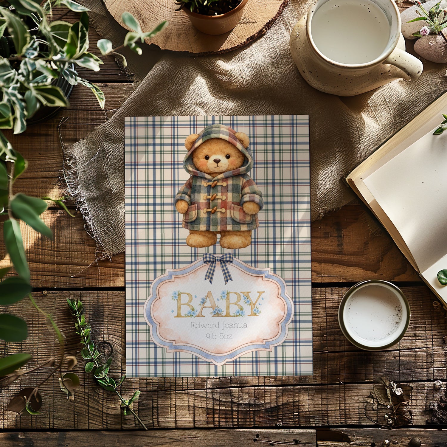 A baby shower card featuring a teddy bear in a plaid hooded jacket, styled on a rustic wooden table with a mug, book, and plants.