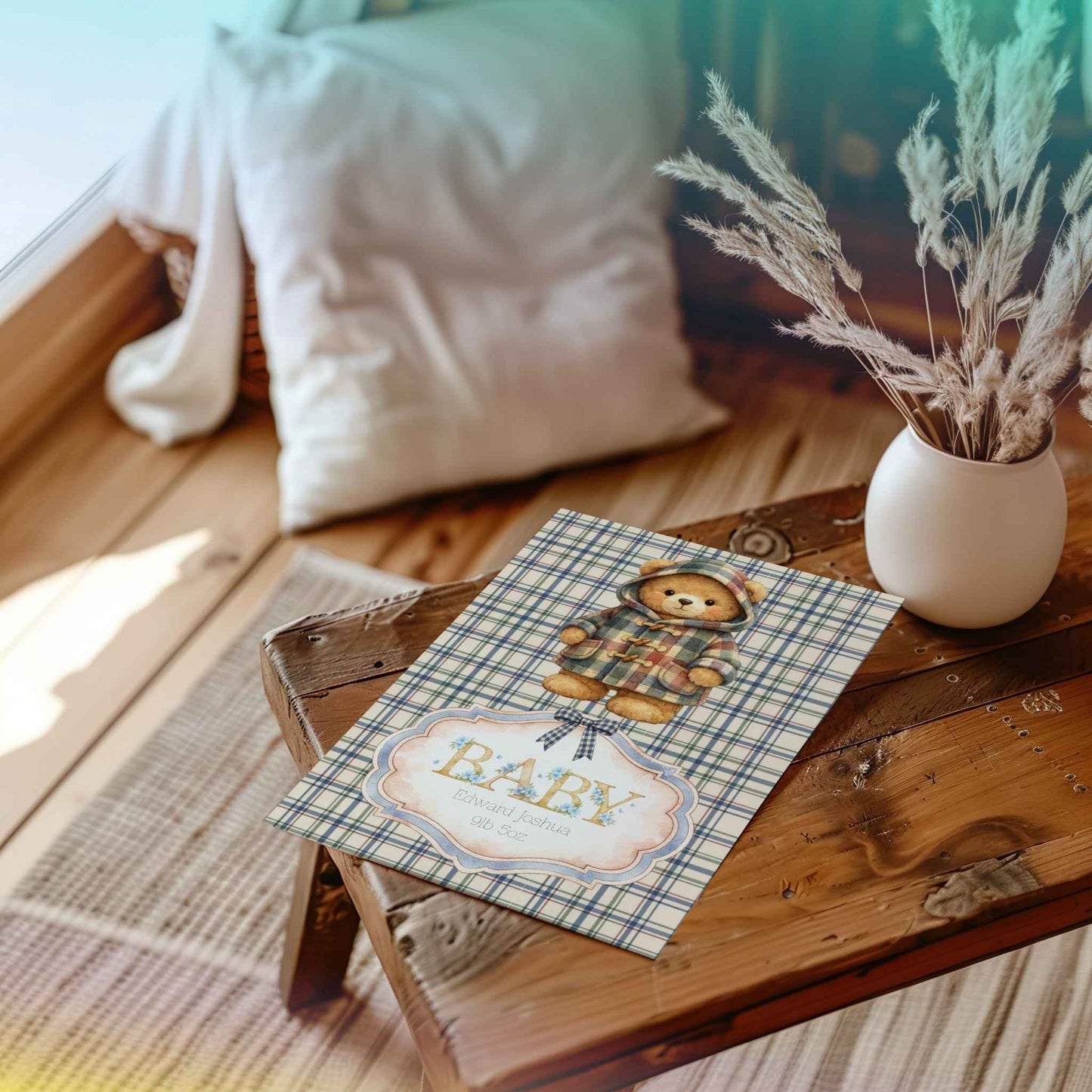 A baby shower card featuring a teddy bear in a plaid outfit and the word 'BABY' on a plaid background, styled on a rustic wooden table with a vase of dried grass and a pillow in the background.