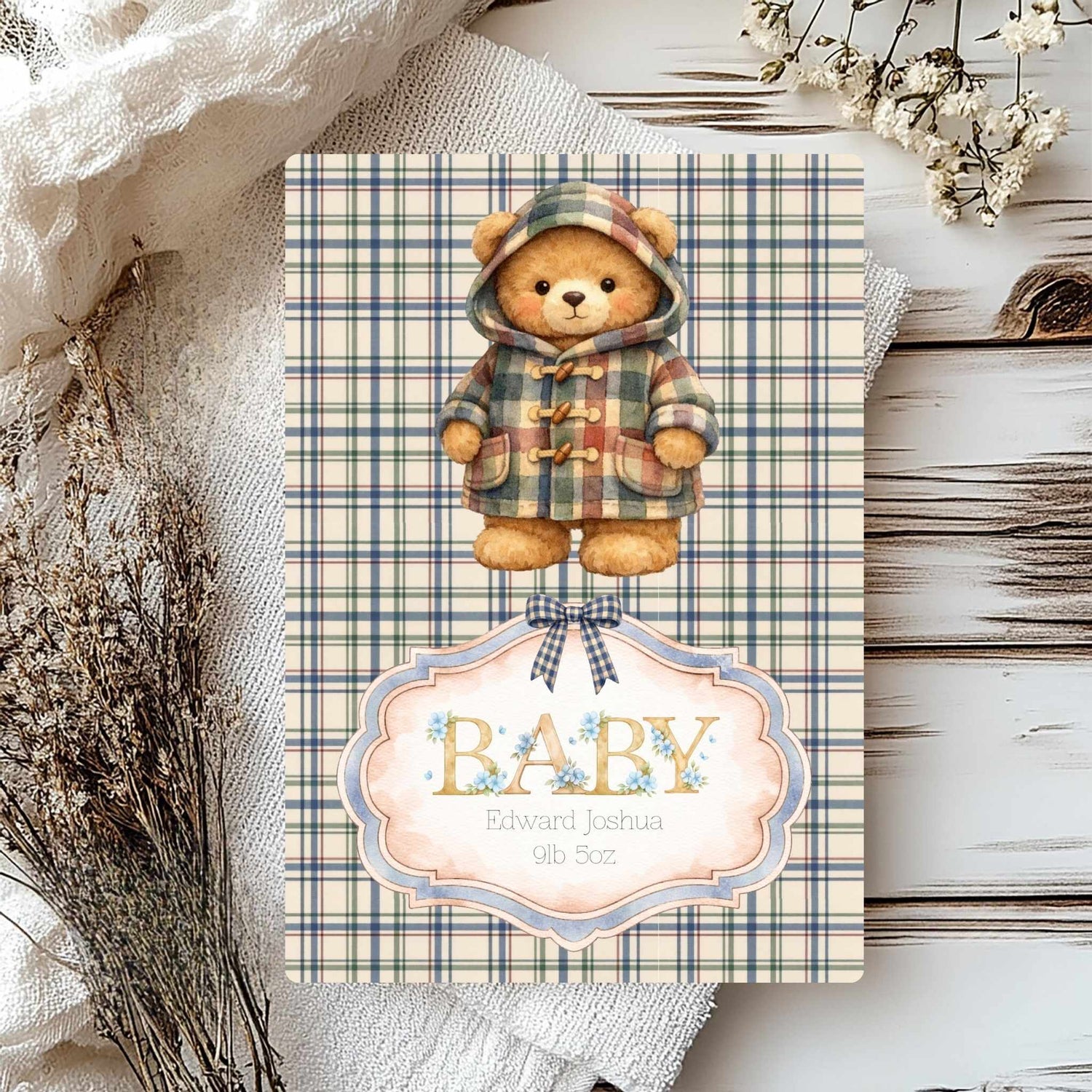 A baby shower card featuring a teddy bear in a plaid hooded jacket, with a decorative label reading 'BABY' and the name 'Edward Joshua 9lb 5oz'. The card is styled on a rustic white wood surface with linen fabric and dried flowers.