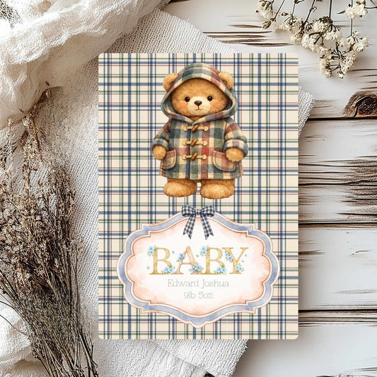 A baby shower card featuring a teddy bear in a plaid hooded jacket, with a decorative label reading 'BABY' and the name 'Edward Joshua 9lb 5oz'. The card is styled on a rustic white wood surface with linen fabric and dried flowers.