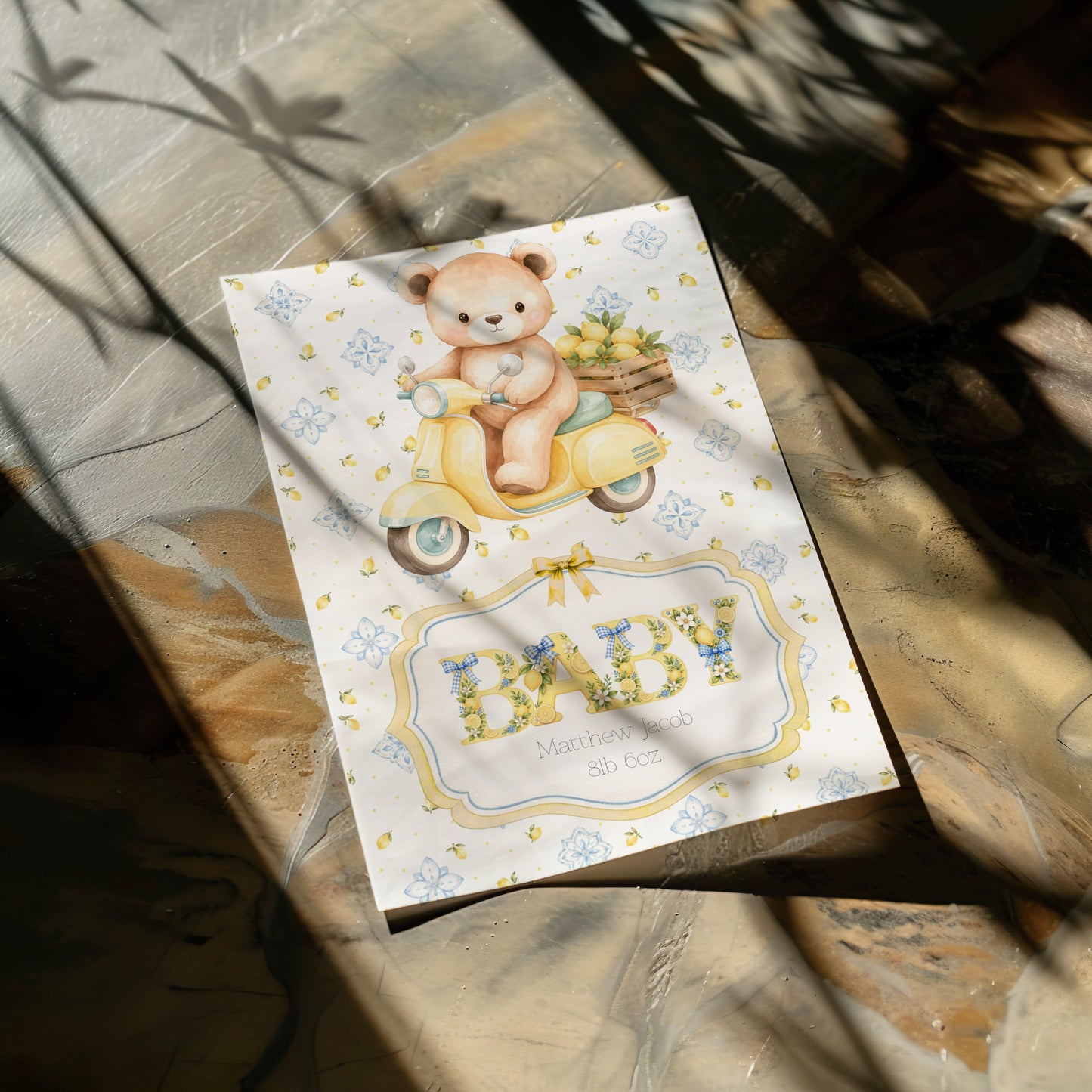 A baby card featuring a cute illustration of a teddy bear riding a yellow scooter and the word 'BABY' in floral letters, photographed on a stone surface with dappled sunlight and shadows.