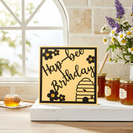 A square greeting card with a yellow background and black border, featuring the words 'Hap-bee Birthday' in script with black flowers and a beehive illustration. The card is styled on a white tray on a wooden table, surrounded by honey jars, a honey dipper, and a vase of white daisies and purple flowers in front of a stone wall and window.