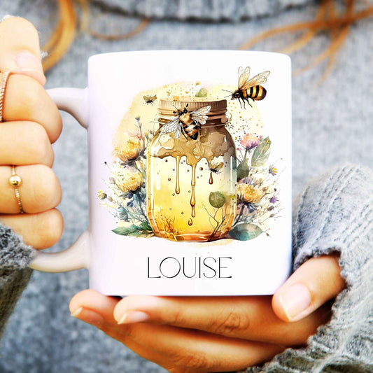 Person holding a mug with a honey jar and bee design, personalised with 'LOUISE'.