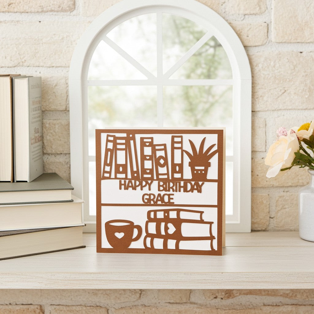 Decorative birthday card with books and a cup on a wooden surface with books and a vase in the background.