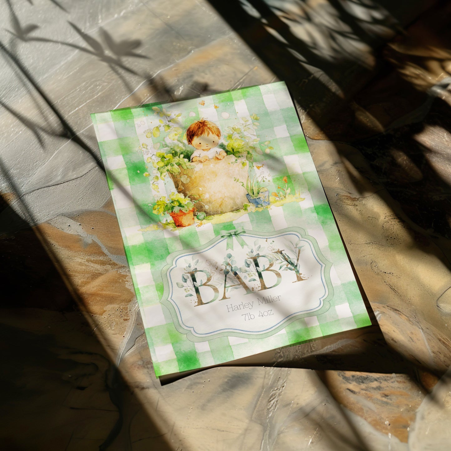 A baby card featuring a green and white gingham pattern, a cute illustration of a baby in a bathtub, and the word 'BABY' photographed on a stone surface with dappled sunlight and shadows.