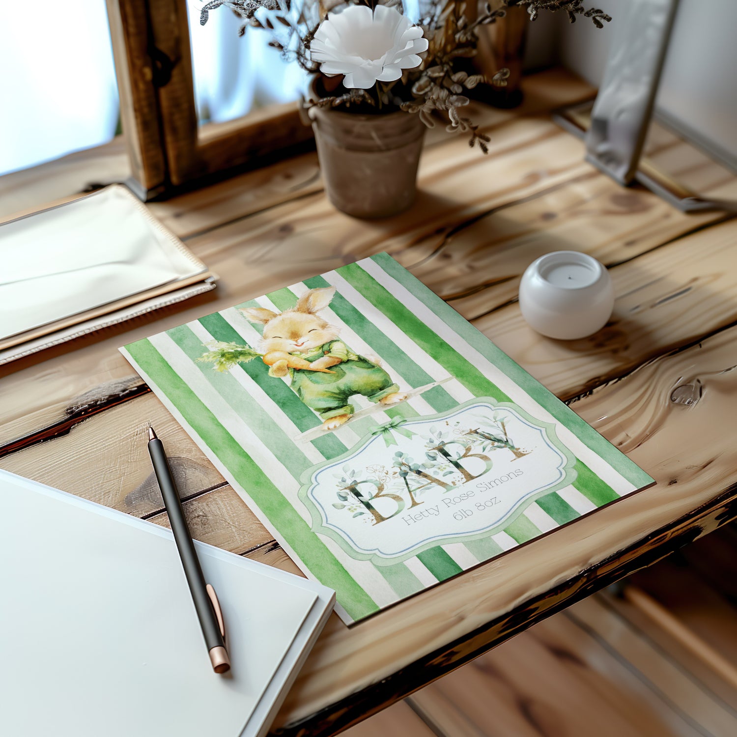 A baby card with green and white stripes, a bunny illustration, and the word 'BABY', styled on a rustic wooden table with a pen, notebook, candle, and dried flowers in the background.