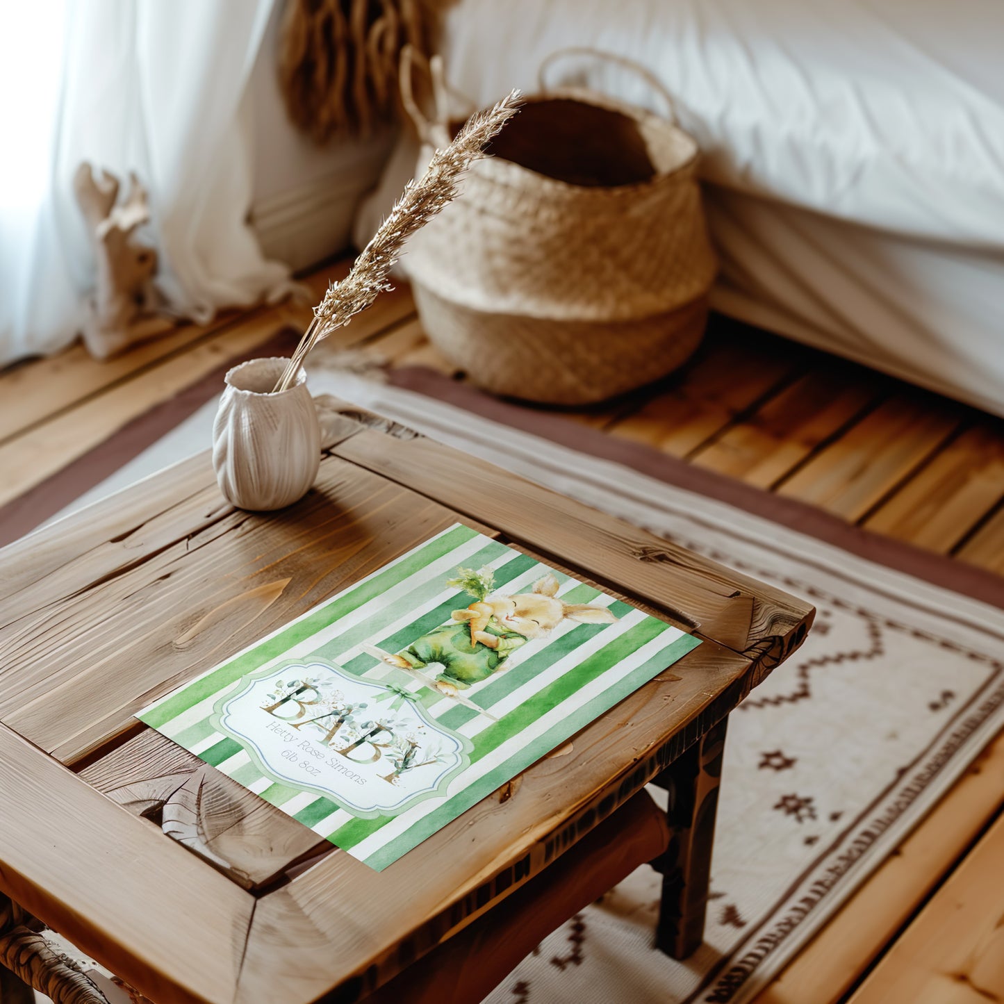 A baby shower card with green and white stripes and a cute bunny illustration sits on a rustic wooden table in a cosy, boho-style nursery setting with a woven basket and dried wheat stem.