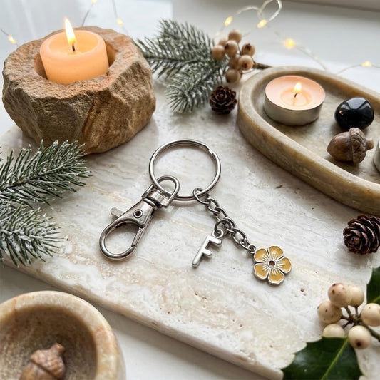 A silver-toned metal keychain with a lobster clasp, a small key charm, and a yellow flower charm, displayed on a light stone tray surrounded by candles, pine branches, acorns, and string lights.
