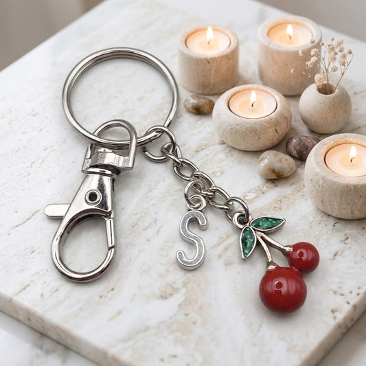 A silver-toned metal keychain with a lobster clasp, a hanging letter 'S' charm, and a cherry charm with red berries and green leaves, displayed on a light marble surface with lit candles in the background.