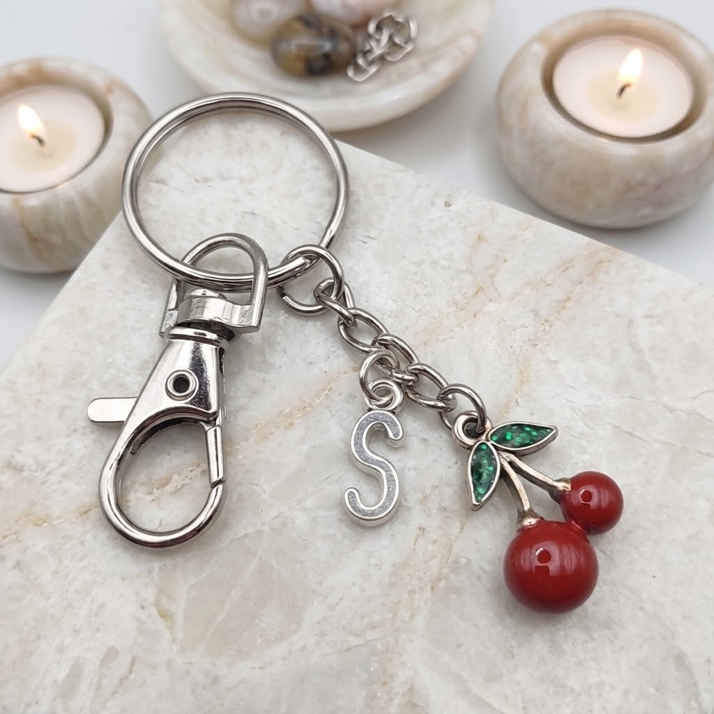 A silver metal keychain with a lobster clasp, a hanging letter 'S' charm, and a cherry charm with red cherries and green leaves, displayed on a light stone slab with blurred lit candles in the background.