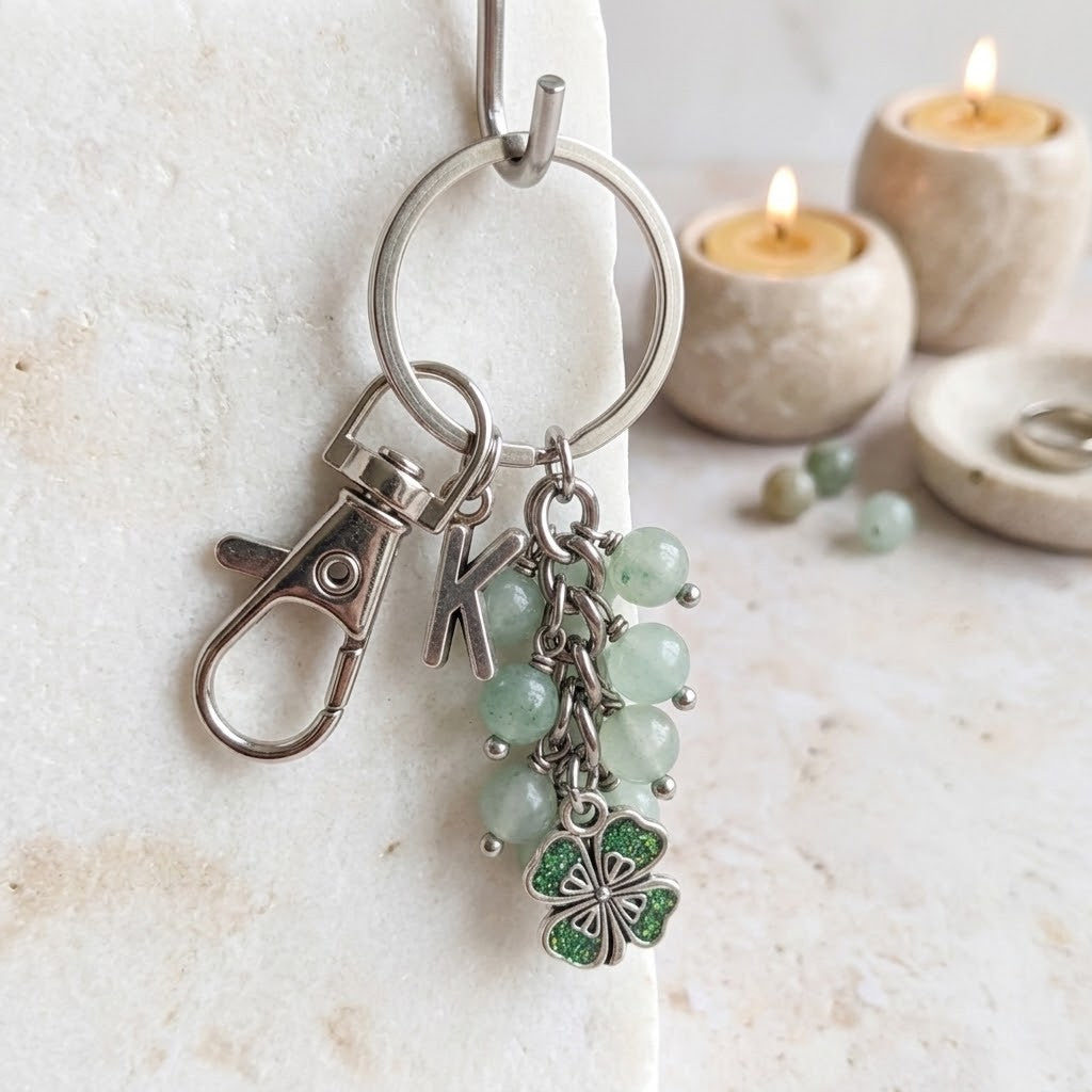 A silver-tone keychain with a lobster clasp, a letter K charm, light green bead strands, and a green four-leaf clover charm, displayed on a textured white surface with blurred candles in the background.