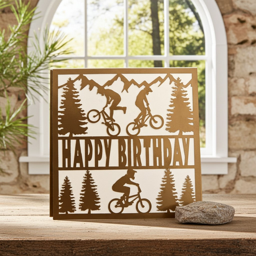A square birthday card with a mountain bike theme, featuring silhouettes of riders, pine trees, and the words 'HAPPY BIRTHDAY' in a warm metallic brown colour, displayed on a wooden surface in front of a rustic stone wall and arched window.