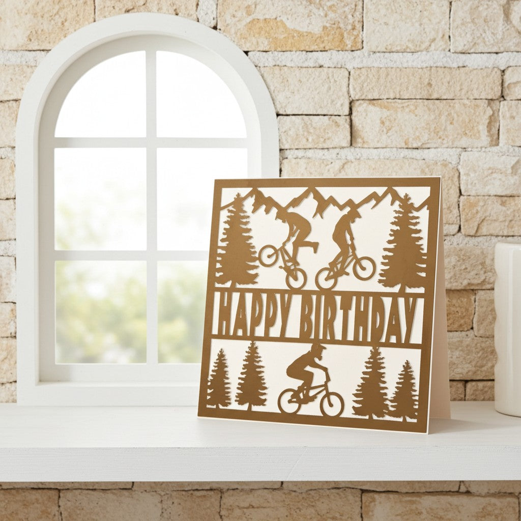 A rectangular brown cardboard birthday card with mountain biker and pine tree silhouettes, standing on a white windowsill against a beige stone wall.