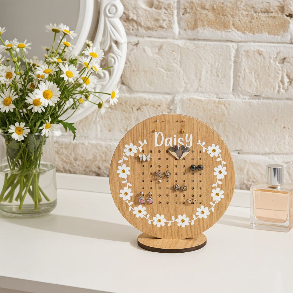 A round wooden jewelry display stand engraved with the name 'Daisy' and decorated with a white daisy floral border, holding several pairs of earrings on a white tabletop. A vase of daisies, a white mirror, and a perfume bottle are visible in the softly lit background.