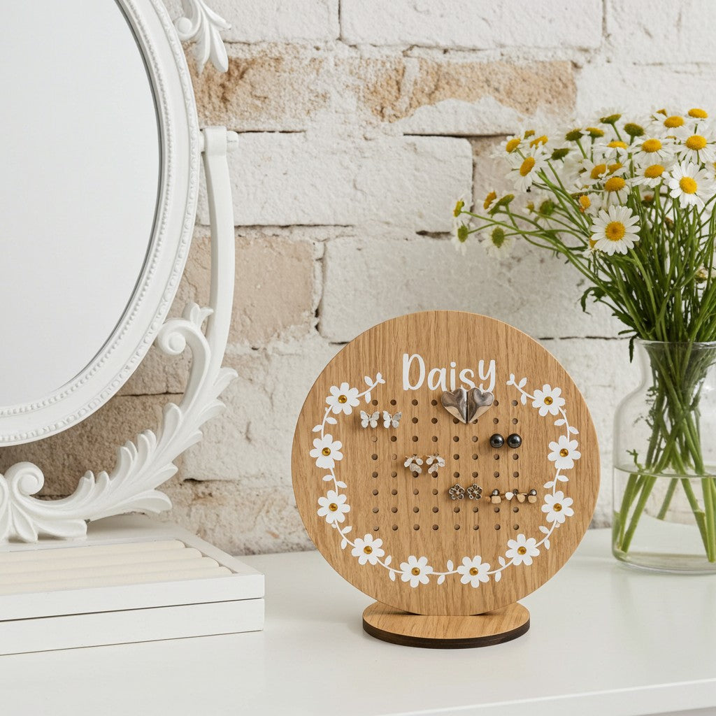 A round wooden jewelry display stand with a stand-up base, featuring a floral daisy border and the name 'Daisy' printed at the top. The stand is styled on a white tabletop next to a white ornate mirror and a glass vase of daisies against a white brick wall.