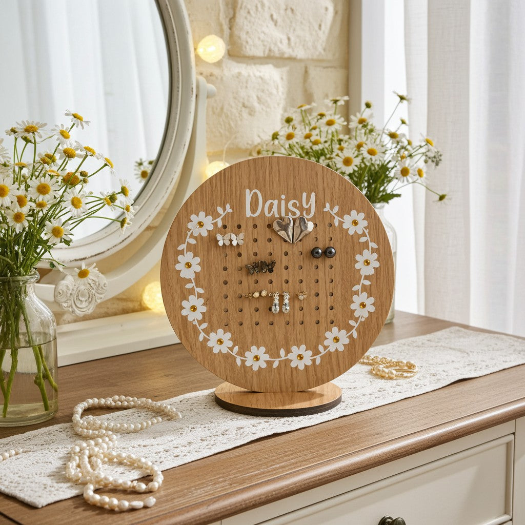 A round wooden jewelry display stand engraved with the name 'Daisy' and decorated with white daisy cutouts around the edge, styled on a wooden dresser with flowers, a mirror, and pearl necklaces.