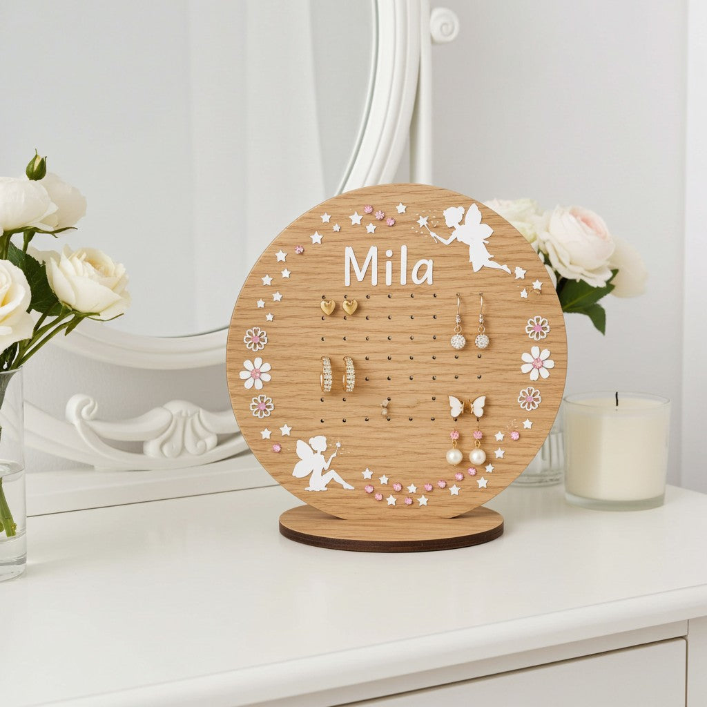 A personalised round wooden jewellery display stand with the name 'Mila' engraved at the top, styled on a white dresser with white roses and a candle in a bright room.