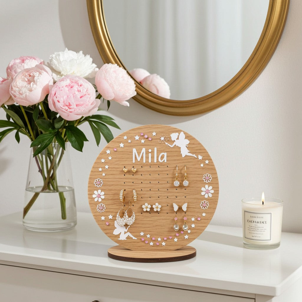 A round wooden jewellery display stand with the name 'Mila' printed on it, styled on a white table next to a vase of pink peonies and a lit candle. The stand features pegs, floral cutouts, and fairy illustrations, holding several pairs of earrings.