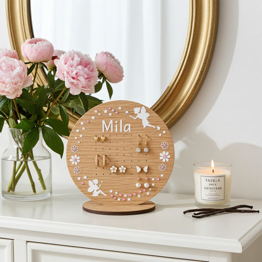 A round wooden jewellery display stand with the name 'Mila' engraved at the top, styled on a white dresser next to a vase of pink peonies, a gold mirror, and a lit candle.