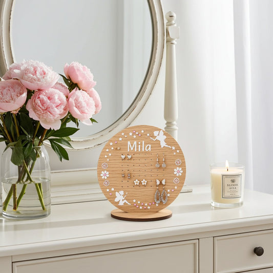 A round wooden jewellery display stand engraved with the name 'Mila' and small angel motifs, styled on a white dresser next to pink peony flowers and a lit candle.