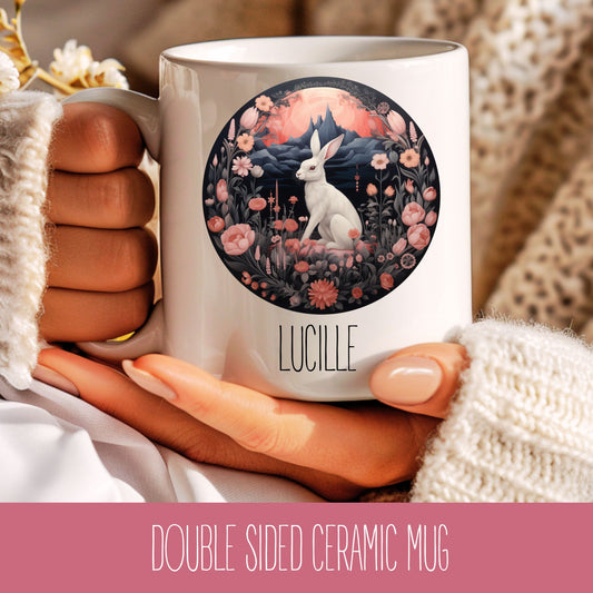A hand holding a white ceramic coffee mug with a floral hare design and the name 'Lucille' printed on it.