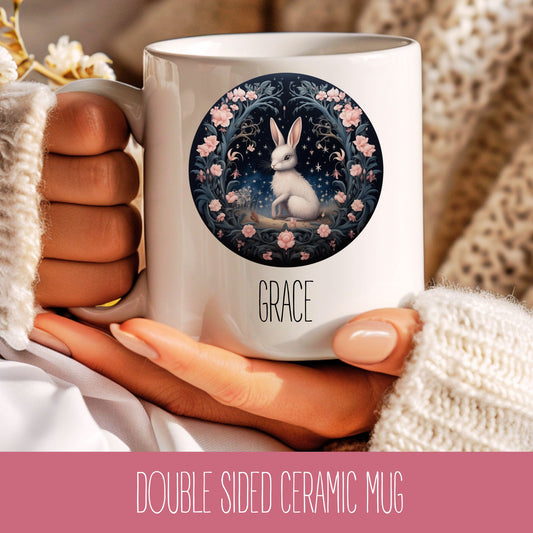 A hand holding a white ceramic mug with a floral hare motif and the name 'Grace' printed on it.
