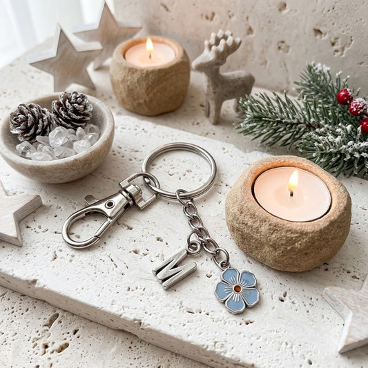 A silver metal keychain with a lobster clasp, a letter charm, and a light blue flower charm with a red centre, displayed on a textured stone surface with candles, pine branches, and decorative stars.