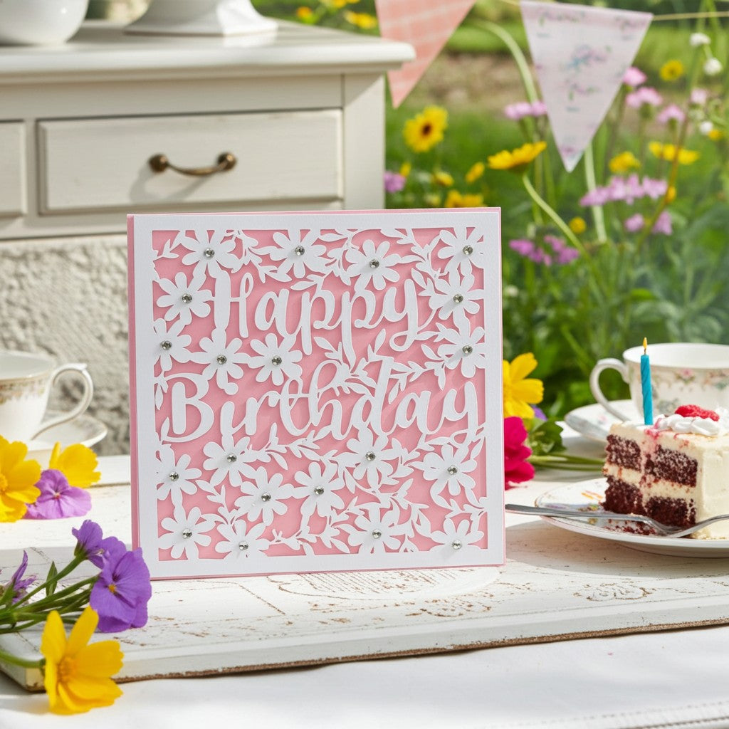 A square pink and white laser-cut birthday card with 'Happy Birthday' in script and floral cutouts, displayed on a table beside a slice of cake, teacups, and flowers in a garden setting.