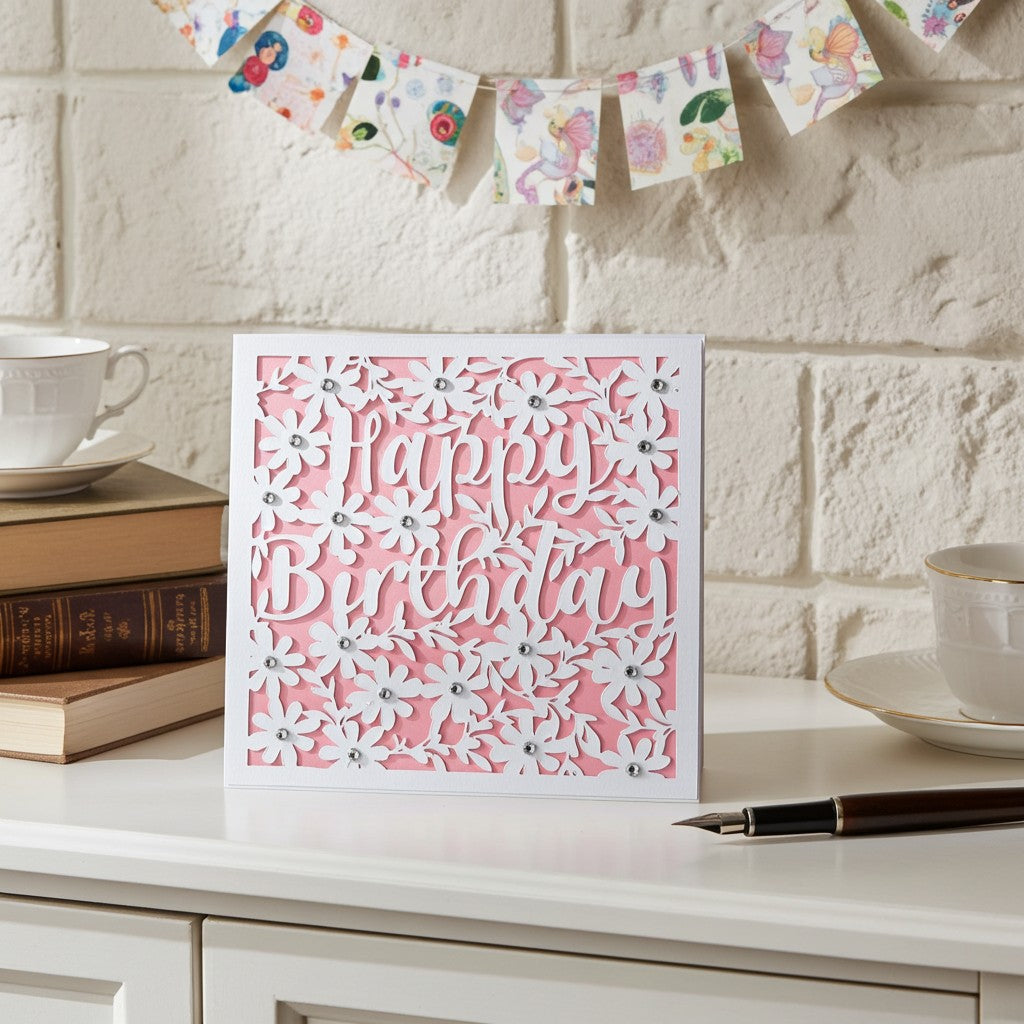 A square white greeting card with layered white floral cutouts and the words 'Happy Birthday' in white script over a pink background, displayed on a white tabletop with books, a teacup, and a pen against a white brick wall.
