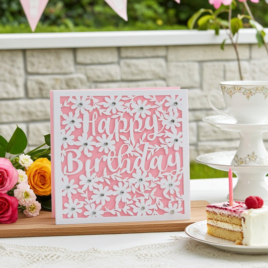A pink and white laser-cut 'Happy Birthday' greeting card displayed on a wooden board in a styled garden party setting with flowers, a cake slice, and a teacup on a pedestal.