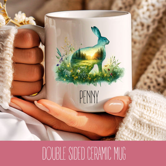 A hand holding a double-sided ceramic mug with a rabbit design and the name 'Penny' on it.