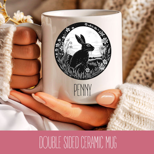 A white ceramic coffee mug with a double-sided design featuring a hare (rabbit) and floral elements, and the name 'Penny' written on it.