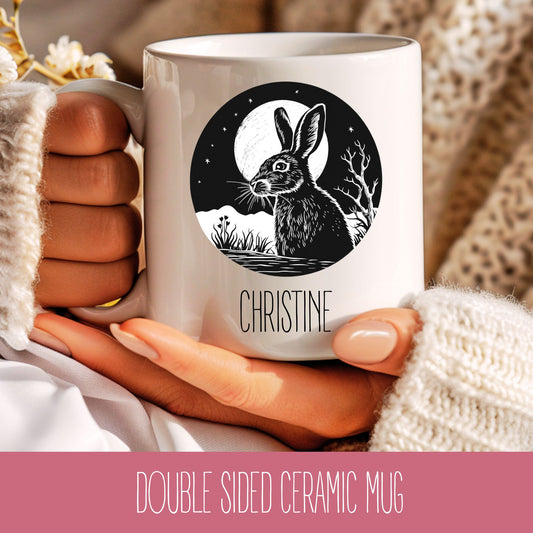 A hand holding a white ceramic mug with a double-sided design featuring a black and white illustration of a hare (rabbit) with a moon and stars in the background.