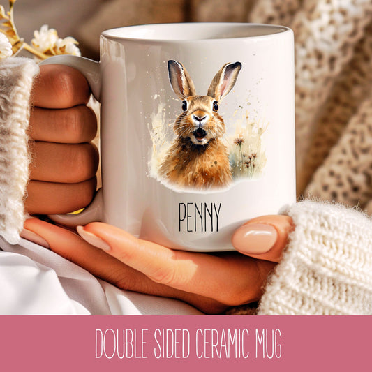 A double-sided ceramic coffee mug with an illustration of a hare on it, held in a hand with a knitted sweater and a cozy in the background.