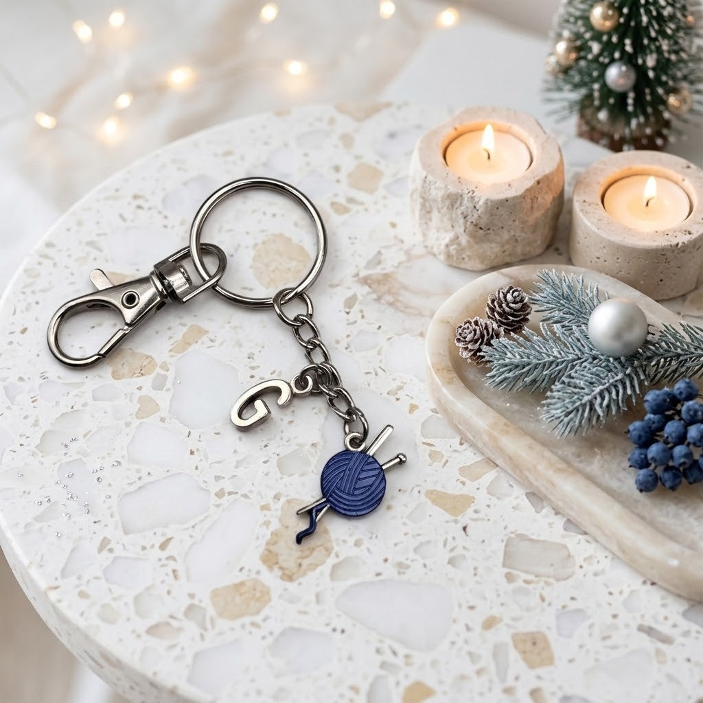 A silver-toned metal keychain with a lobster clasp, key ring, and dangling charm shaped like blue yarn ball and knitting needles, displayed on a speckled white stone tray with candles, pinecones, and a small tree in the background.