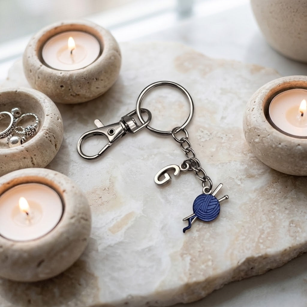 A silver-toned metal keychain with a lobster clasp, key ring, and dangling charm shaped like a blue yarn ball with knitting needles, displayed on a stone slab with lit tealights in the background.