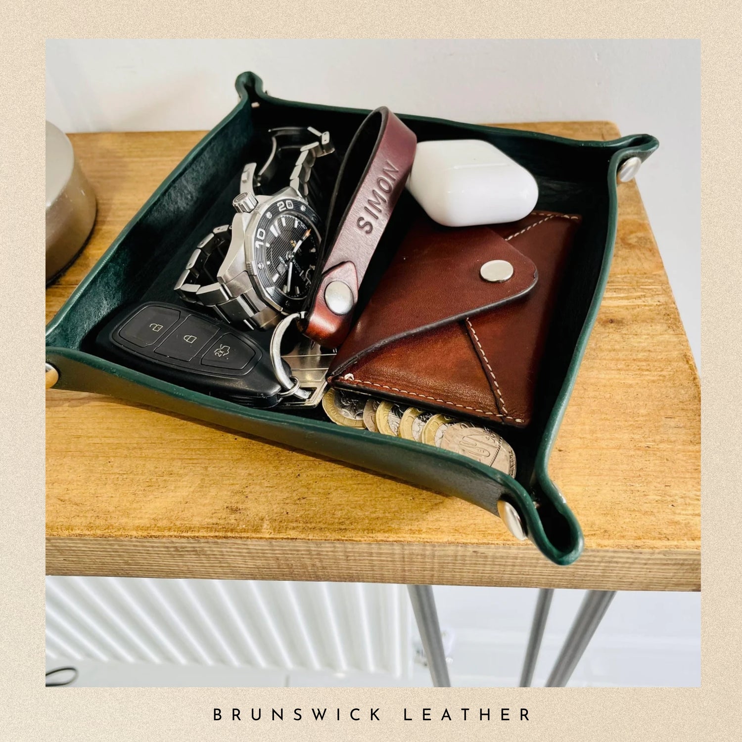 Green leather tray with a watch, keys, phone case, and wallet on a wooden surface, branded 'Brunswick Leather'.