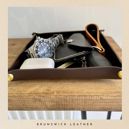 Brown leather tray with watch, wallet, and keychain on a wooden surface, branded 'Brunswick Leather'.