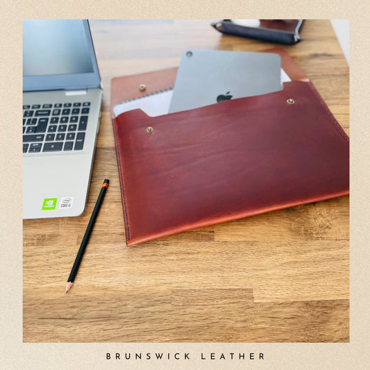 Brown leather laptop sleeve with Apple product on a wooden surface, branded 'Brunswick Leather'.