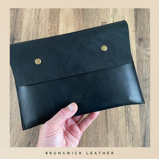 Black leather clutch with gold buttons held by a hand on a wooden surface, branded 'Brunswick Leather'.