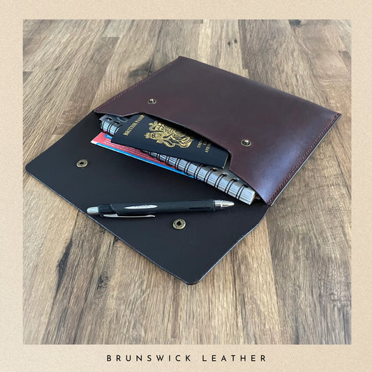Brown leather wallet with passport and pen on a wooden surface, branded 'Brunswick Leather'.