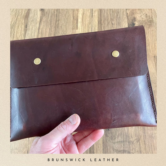 Brown leather clutch held by a hand on a wooden surface with 'Brunswick Leather' branding.