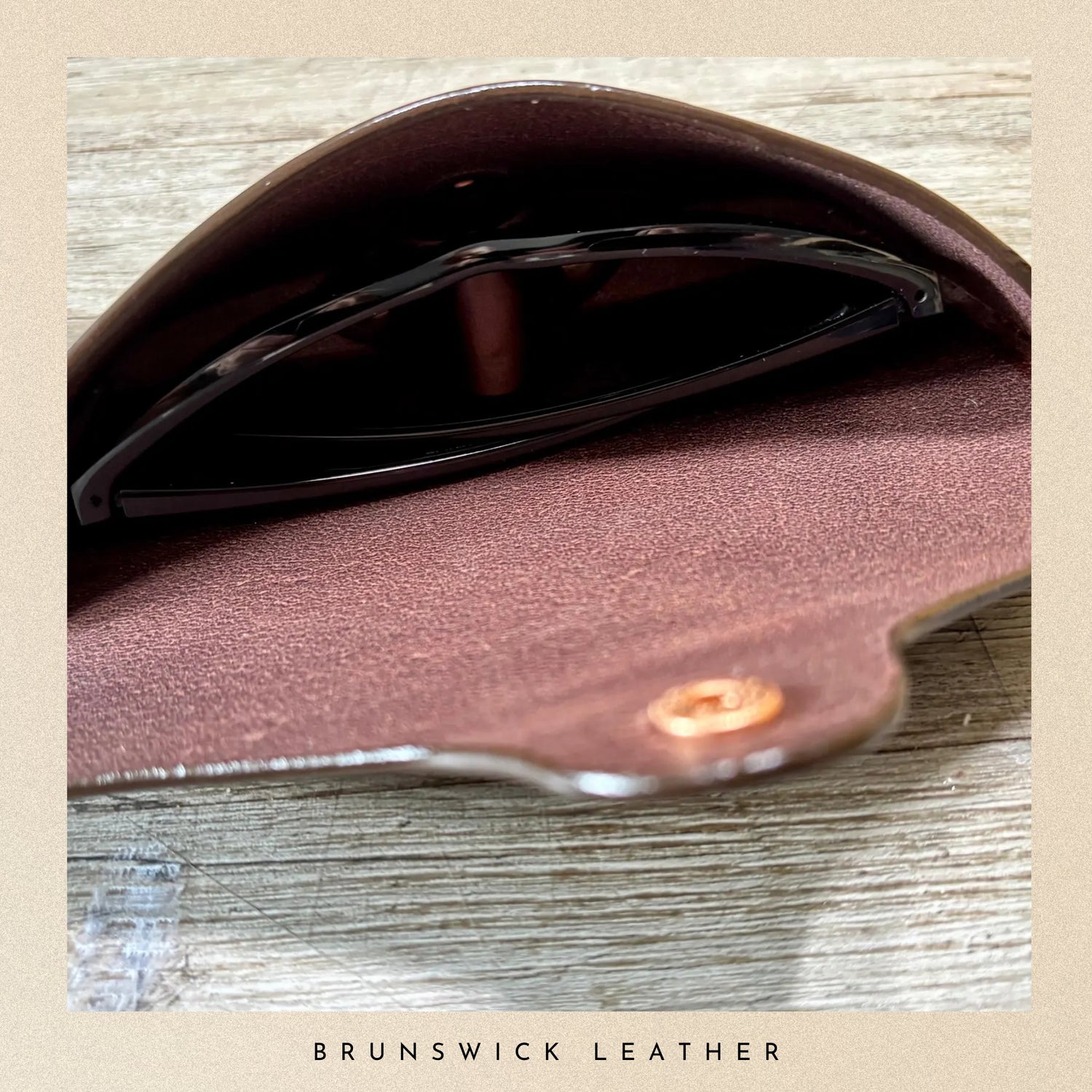 Close-up of a brown leather wallet with sunglasses on a wooden surface, branded 'Brunswick Leather'.