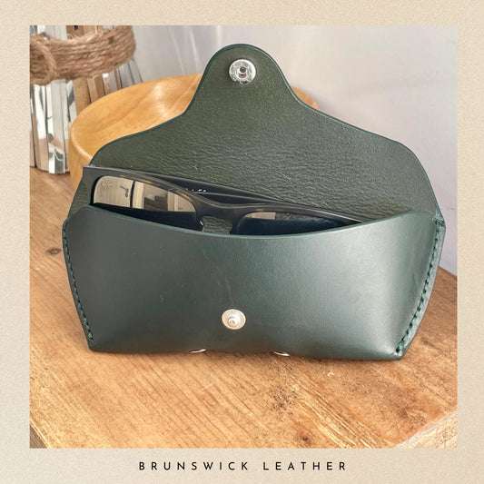 Green leather case for sunglasses on a wooden surface with 'Brunswick Leather' branding.